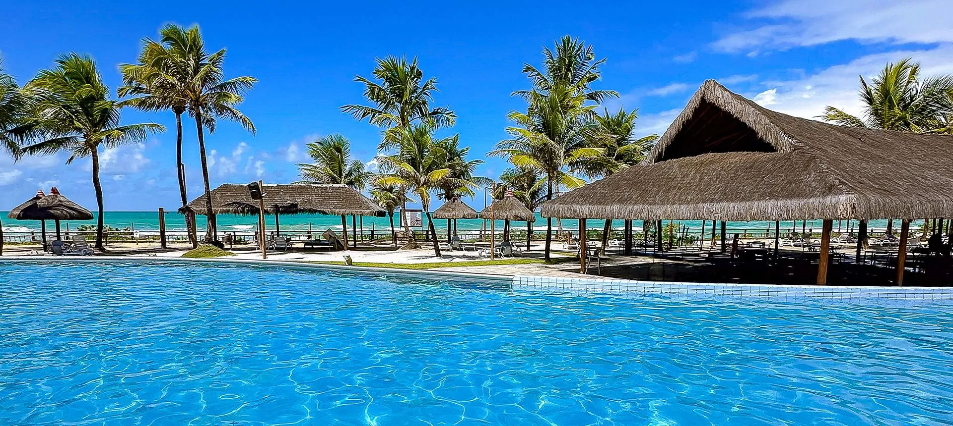 Área de descanso com espreguiçadeiras e quiosques de palha integrados à grande piscina azul do resort Enotel Porto de Galinhas
